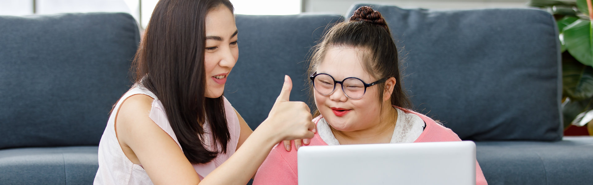 mother sitting on sofa in living room teaching a girl with down syndrome kid with eyeglasses using laptop notebook computer