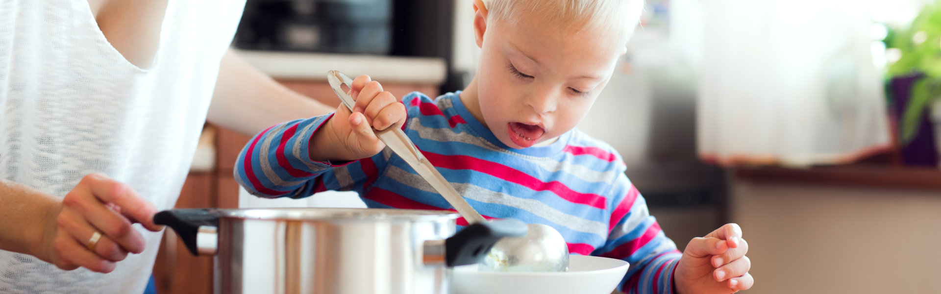 A happy handicapped down syndrome boy with unrecognizable mother pouring soup in a plate indoors